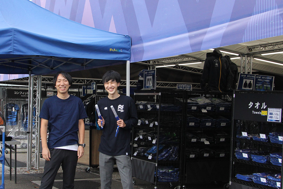 Watanabe'sコラボ藍染Tシャツを着る奥山勇太さん(左)と、PenaltyハイスロングTシャツを着用してペンライトを持つ井苅将樹さん(右)。ホームゲーム会場に設営されたショップの前で